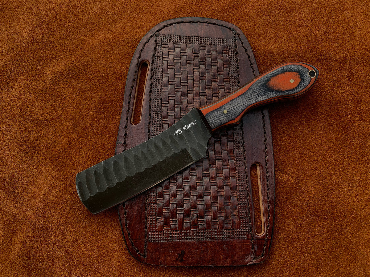1095 steel cowboy bull cutter knife with a black sculpted blade, two-tone orange and charcoal Pakka wood handle with brass pin, resting on a basket-weave tooled leather sheath