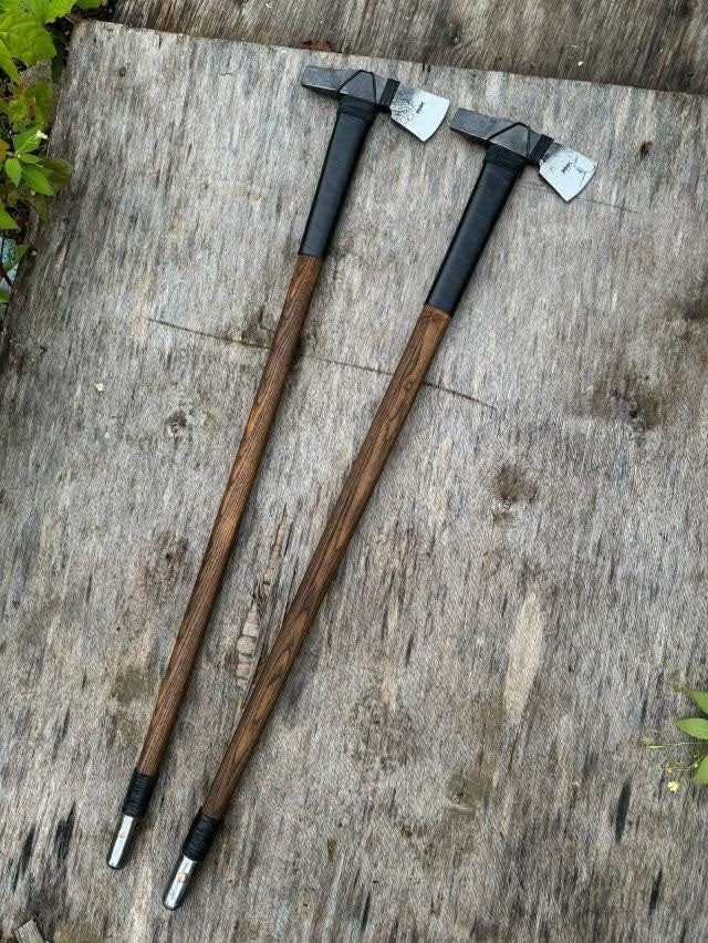Two long-handled Axe with brown wooden handles, black grip sections, and dark metal heads, placed on a weathered gray wooden surface.