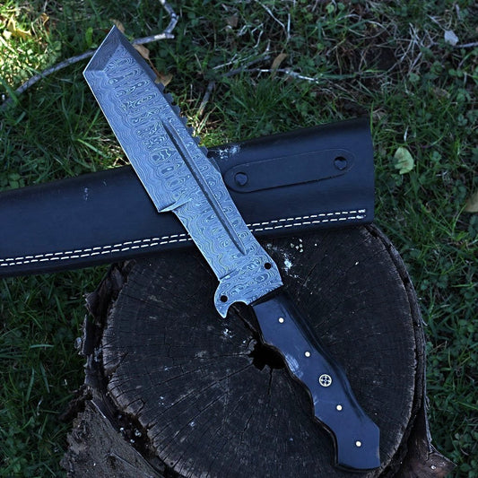 A custom handmade Damascus tracker knife with a black horn handle and a leather sheath, displayed on a wooden stump with grass and trees in the background.