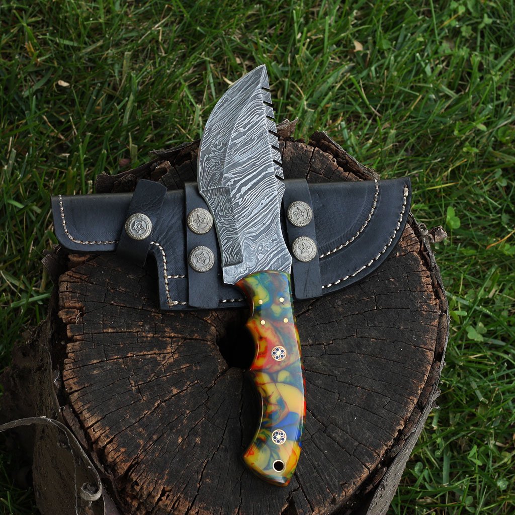 A handmade Damascus steel knife with a multicolored handle and a black leather sheath, resting on a wooden stump with grass and bushes in the background.