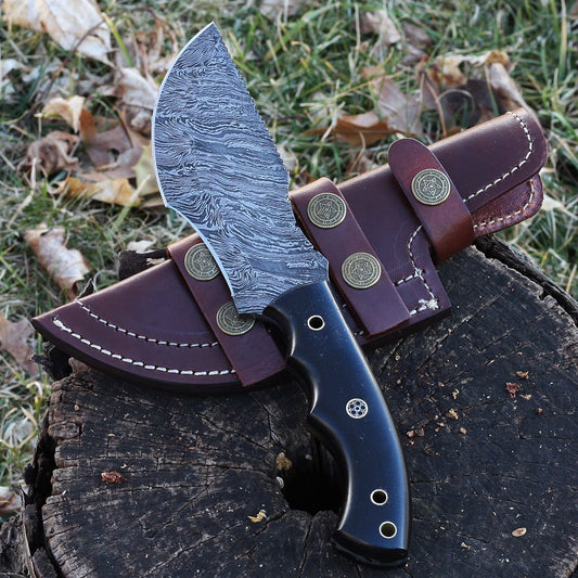 A knife with a Damascus pattern blade and a brown leather sheath, resting on a wooden surface with fallen leaves around.