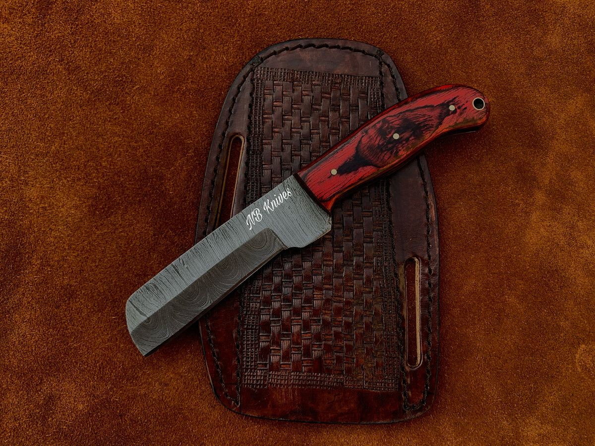 A custom handmade Damascus cowboy bull cutter knife with a black and red handle, placed next to its brown leather sheath on a textured background.